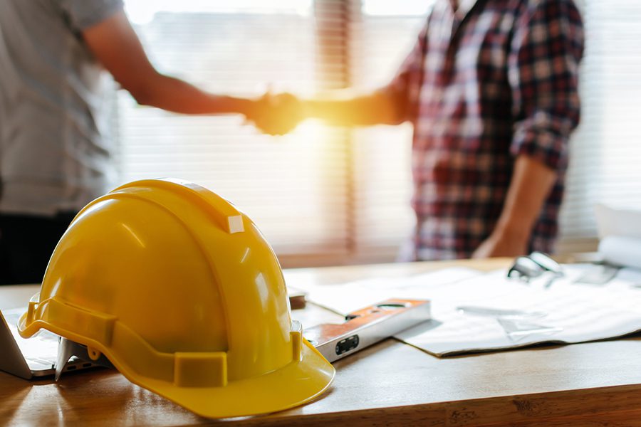 Specialized Business Insurance - Yellow Helmet on Desk with Construction Workers Blurred and Shaking Hands in the Background